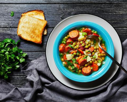 Smoked sausage soup in a bowl on a table with toast fresh herbs and table cloth.